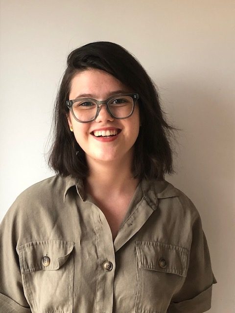 Emma smiling at the camera, wearing a olive green button up and glasses.