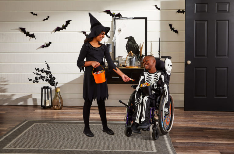 A child dressed as a witch handing candy to a child dressed as a skeleton. He is using a wheelchair.